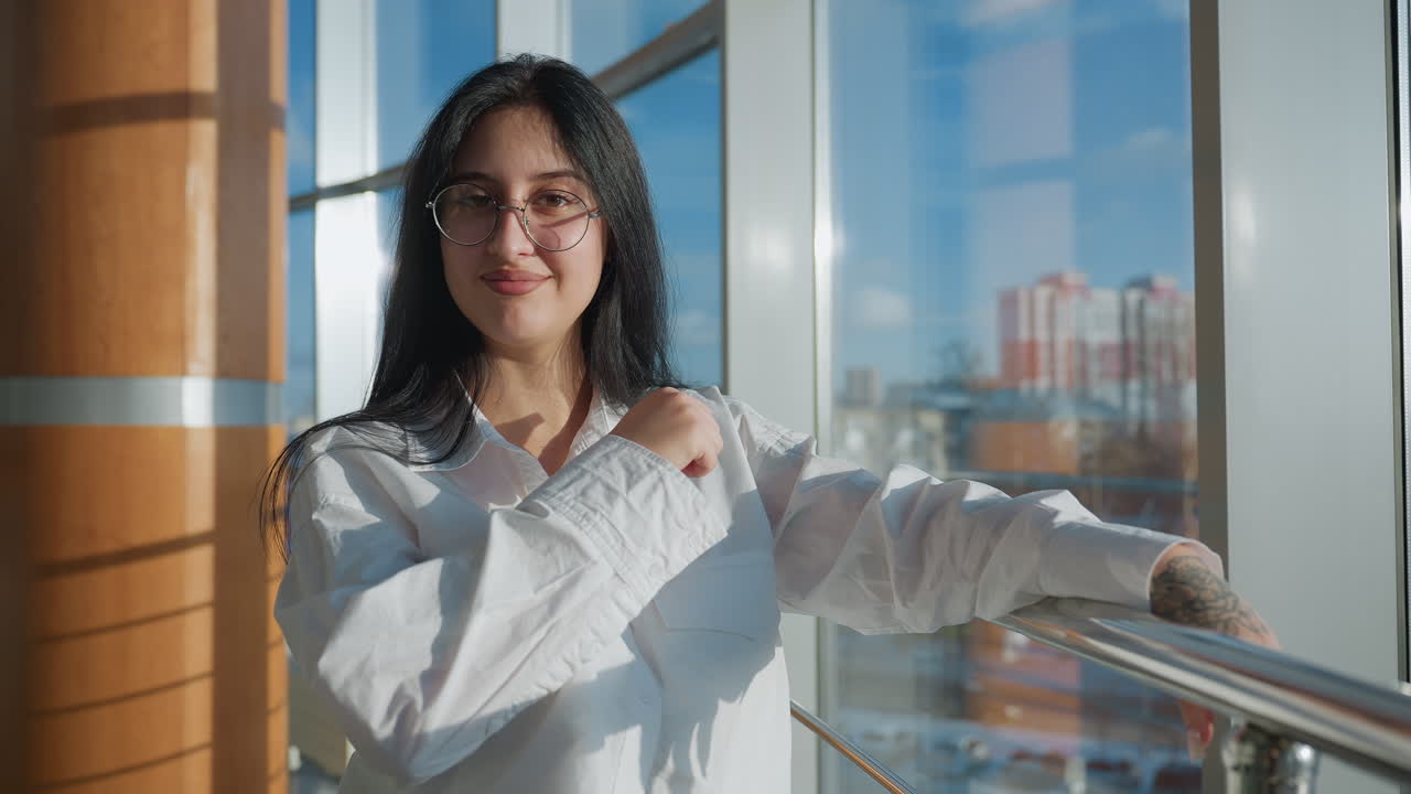 Confident young woman with glasses leaning on railing and smiling while making playful hand gesture in brightly lit modern space, sunlight pouring through large windows with urban background view