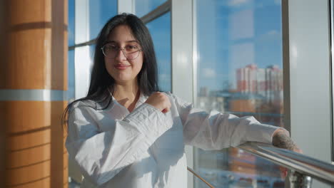 confident young woman with glasses leaning on railing and smiling while making playful hand gesture in brightly lit modern space, sunlight pouring through large windows with urban background view