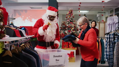 Employee-wearing-Santa-Claus-costume-collecting-unneeded-clothes-from-generous-shoppers-in-donation-container,-giving-them-as-gift-during-Christmas-season-charity-efforts,-spreading-holiday-cheer