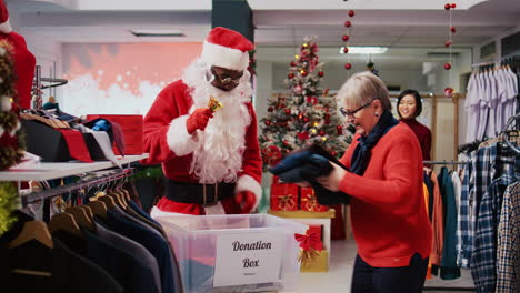 employee wearing santa claus costume collecting unneeded clothes from generous shoppers in donation container, giving them as gift during christmas season charity efforts, spreading holiday cheer