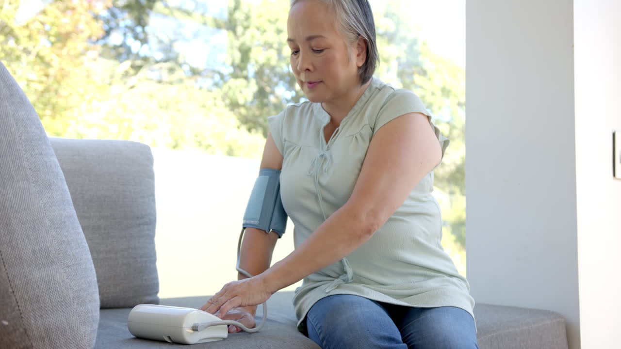 Checking blood pressure, senior asian woman using monitor while sitting on couch
