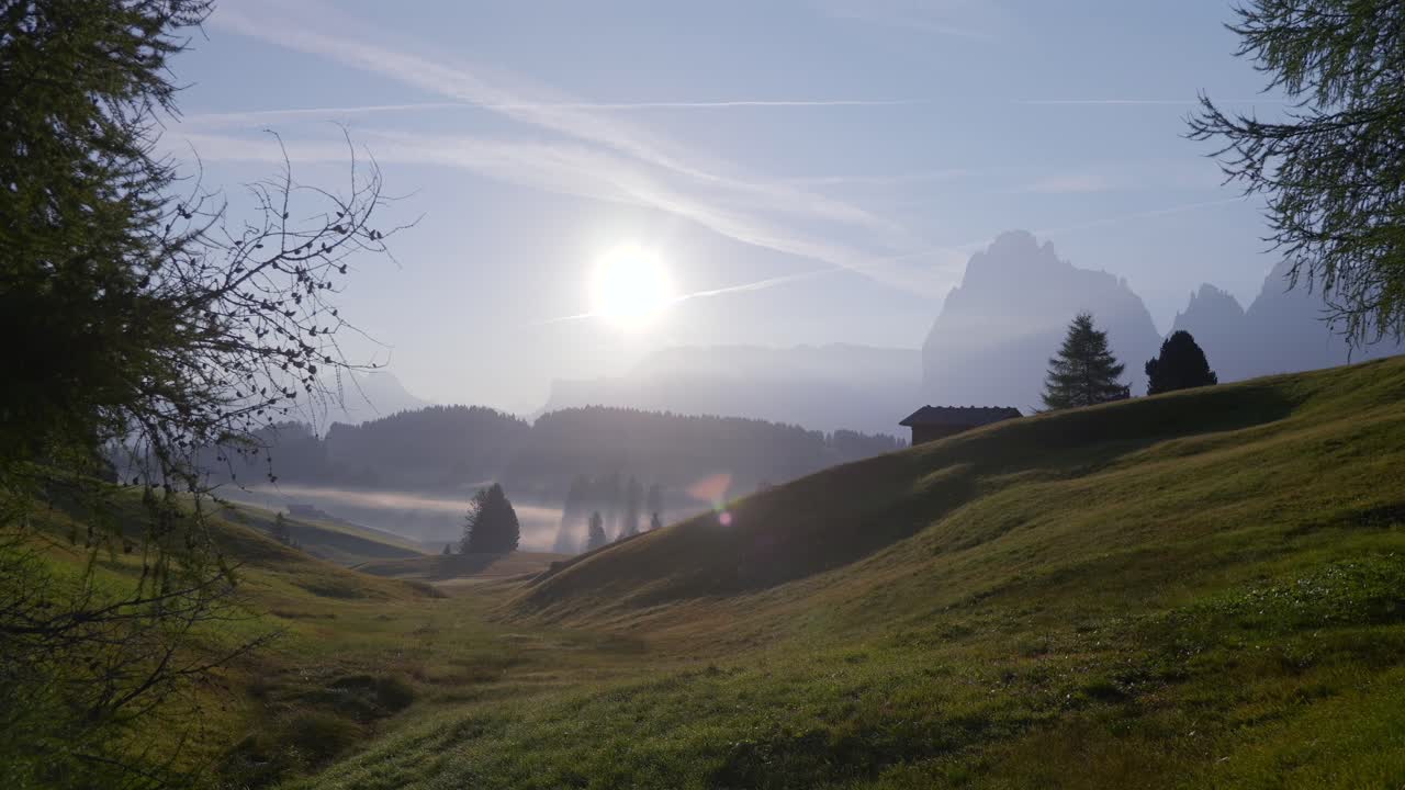 The sun rises over misty meadows and rolling hills in Seiser Alm, casting light through scattered trees with the Dolomite mountains in the background. A serene morning in Italy’s alpine countryside.
