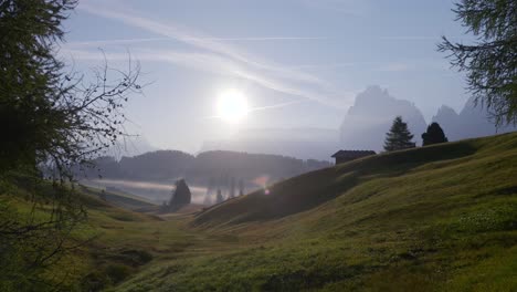 the sun rises over misty meadows and rolling hills in seiser alm, casting light through scattered trees with the dolomite mountains in the background. a serene morning in italy’s alpine countryside.