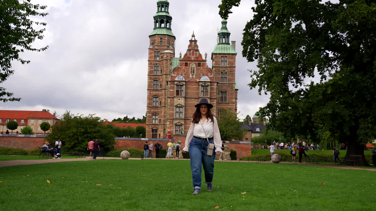 Woman with casual clothes and hat walks towards camera near Rosenborg Castle