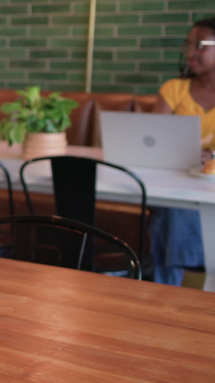 Vertical video: Typing woman in yellow top on laptop, tart at cafe, looking up at passing paper