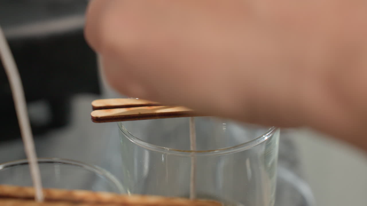 Close up of person adjusting candle wick using wooden clip to keep wick centered inside glass cup during handmade candle making process, showcasing precision and care in craft preparation