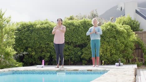 two happy diverse senior women practising yoga by pool in sunny garden, slow motion, copy space