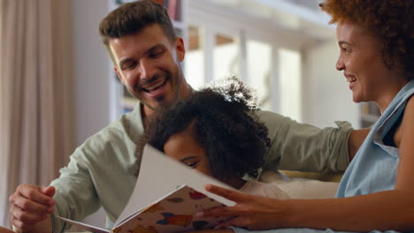 Hija-Leyendo-Un-Libro-Con-Sus-Padres-En-El-Sofá-De-Casa