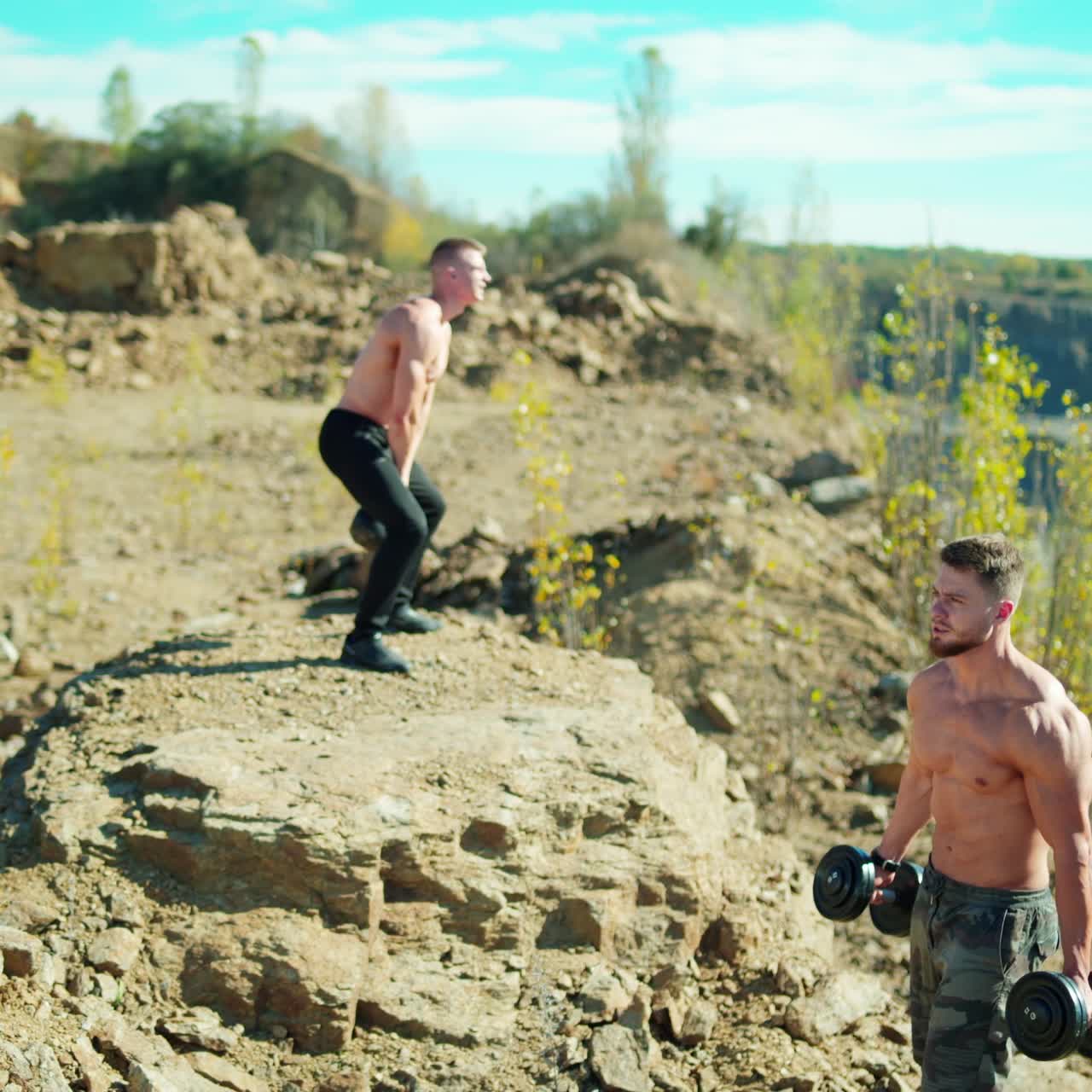Two athletes training with sports equipment. Muscular man lifting dumbbells outdoors. Strong sportsman lifting kettlebell on the rocky hill.
