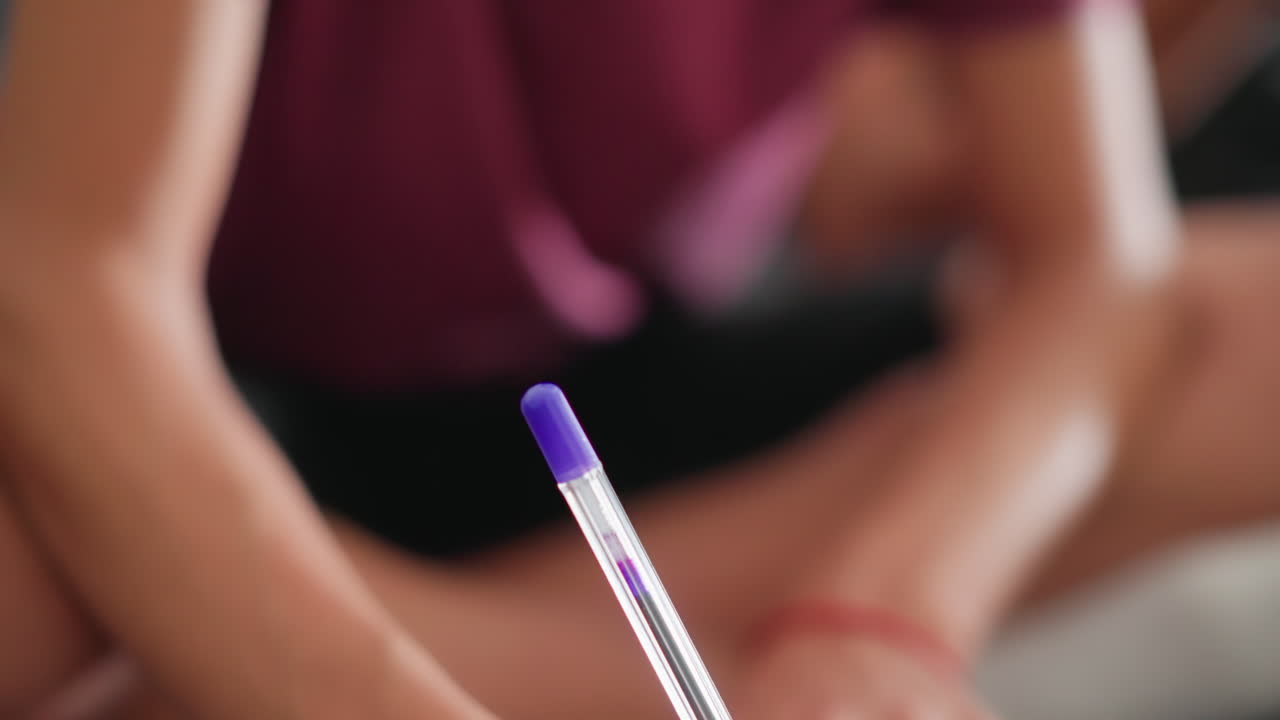 close up of young girl writing with blue pen while seated cross legged on sofa with slightly blurred background showing another person reading nearby