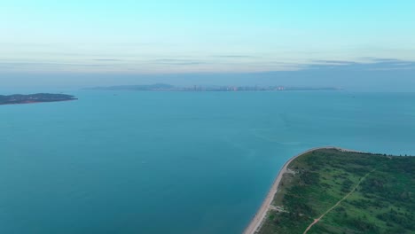 Toma-Aérea-Hacia-Atrás-Que-Muestra-La-Playa-De-Arena-En-La-Isla-Verde-De-Kinmen-金門-Quemoy-Y-La-Ciudad-De-Xiamen-En-El-Fondo-Con-Un-Hermoso-Mar-Durante-El-Día-Brumoso