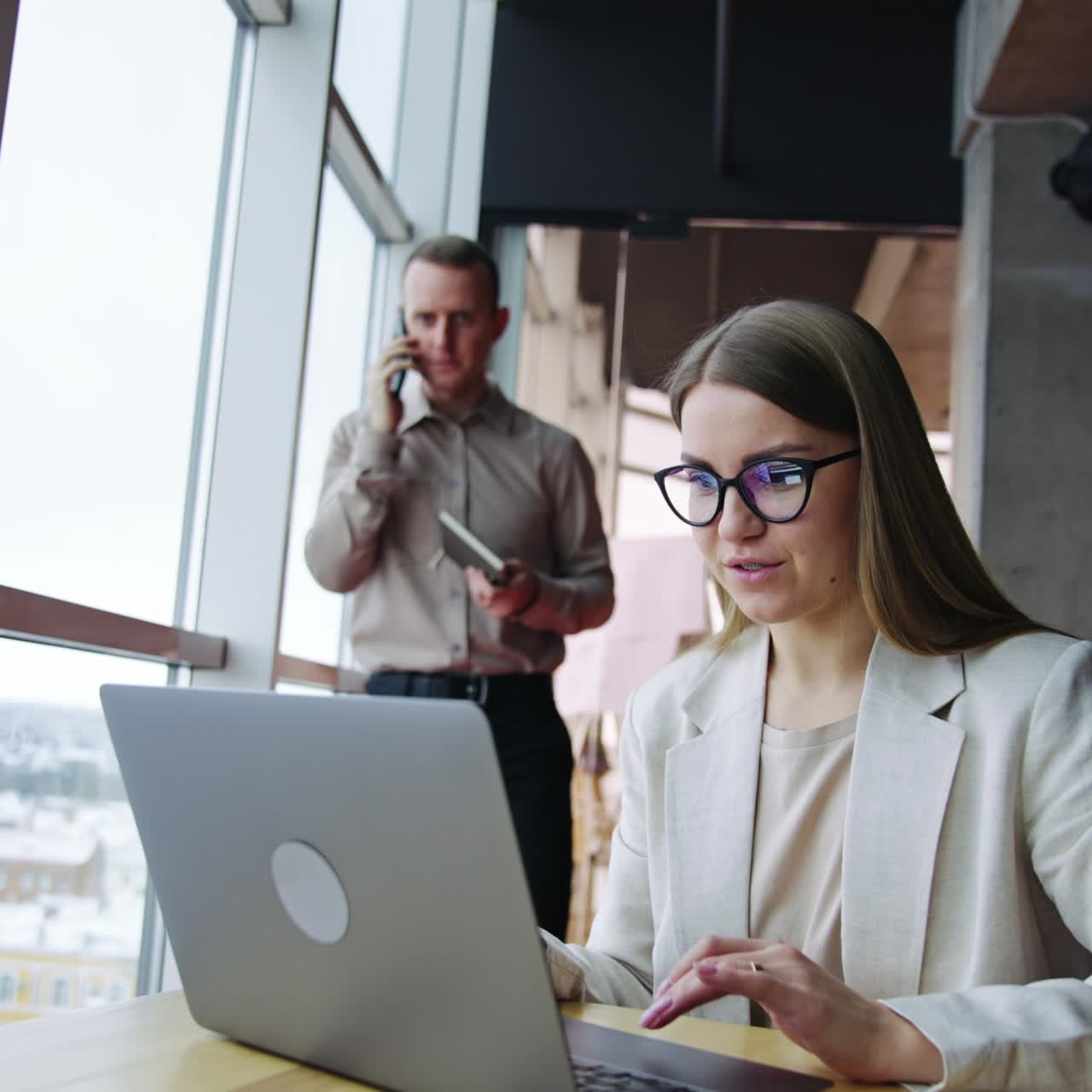 Pleasant-looking woman works on her laptop at table. Man with paper book in his hand talks on the phone at backdrop