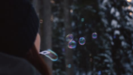 Brünette-Frau-Mit-Winterkleidung,-Die-Regenbogenfarbene-Seifenblasen-Bläst,-Die-In-Der-Luft-Schweben,-Mit-Einem-Verschneiten-Wald-Im-Hintergrund