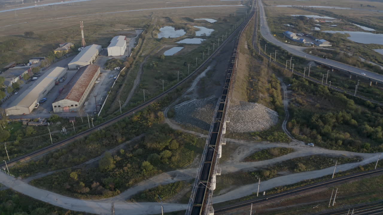 toma aérea de pivote lento de un largo tren de carruajes de carbón completamente cargado, pasando por campos verdes, con almacenes a los lados de la carretera con la revelación de la cresta de la montaña en la lejanía, al atardecer