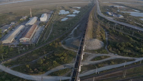 Slow-pivot-aerial-shot-of-a-long-full-loaded-coal-carriage-train,-passing-through-green-fields,-with-warehouses-on-the-road-sides-with-the-reveal-of-mountain-ridge-in-the-far-distance,-on-the-sunset