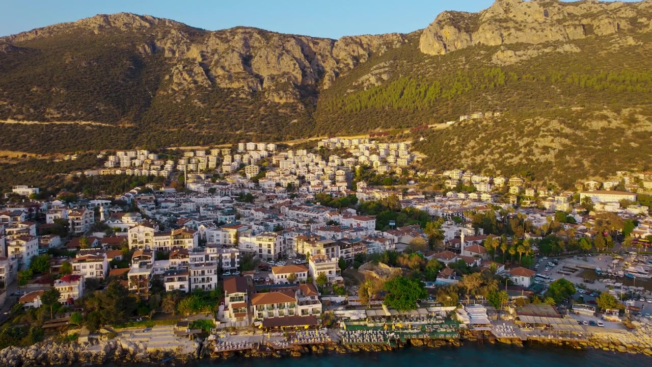 Vista aérea em movimento da cidade costeira de Kas, na Turquia, repleta de edifícios brancos densos, telhados de terracota, estradas sinuosas e minaretes de mesquitas espalhados abaixo de montanhas íngremes de calcário sob a luz quente do dia