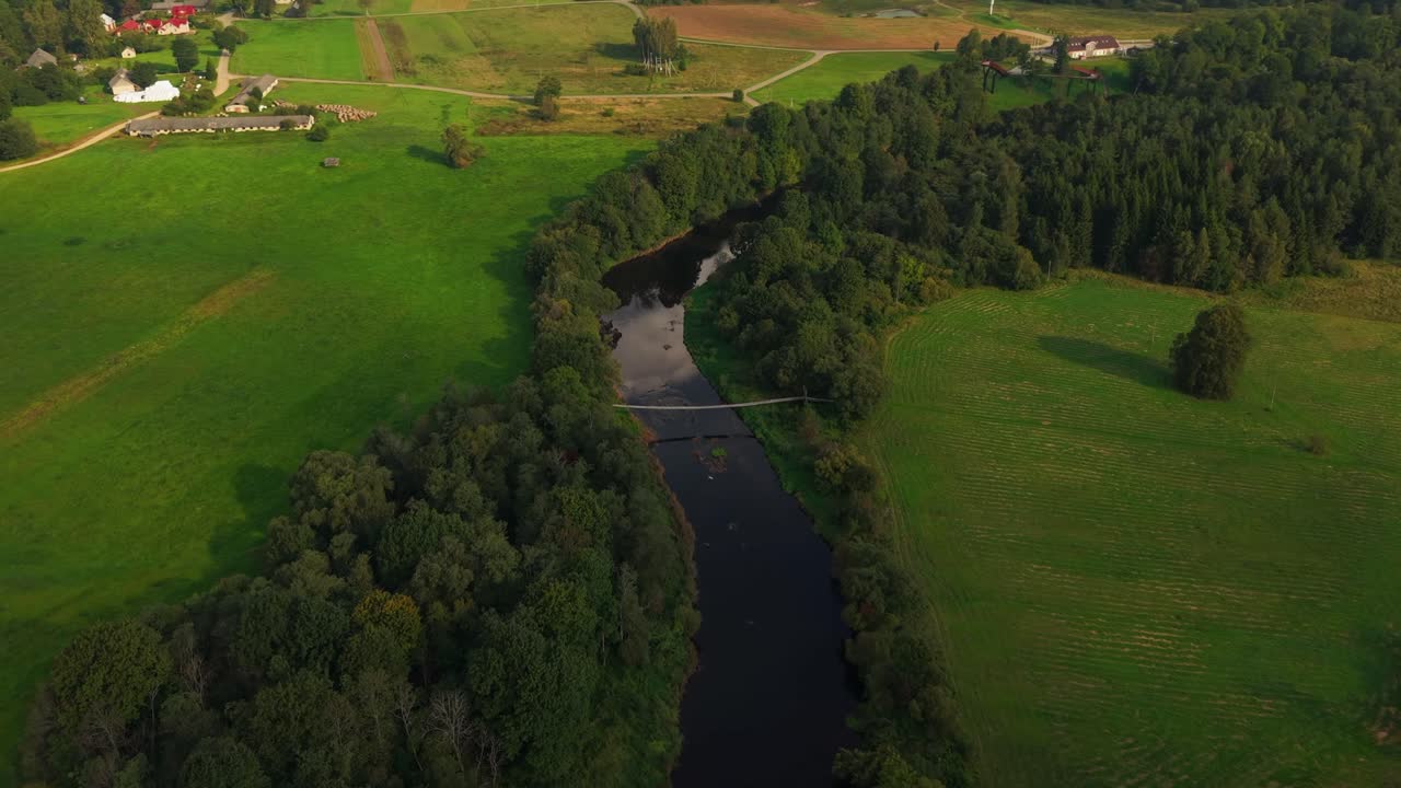 Aerial view of a lush countryside landscape with a winding river, dense forest, open green fields, and a small pedestrian suspension bridge connecting both riverbanks