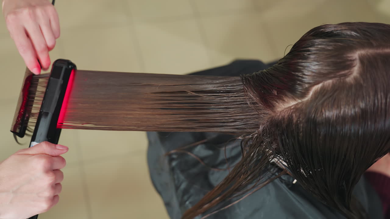 caucasian stylist using flat iron on last hair section to create sleek smooth finish, comb guiding strand, ceramic plates gliding, salon floor tiles visible, professional finalizing step
