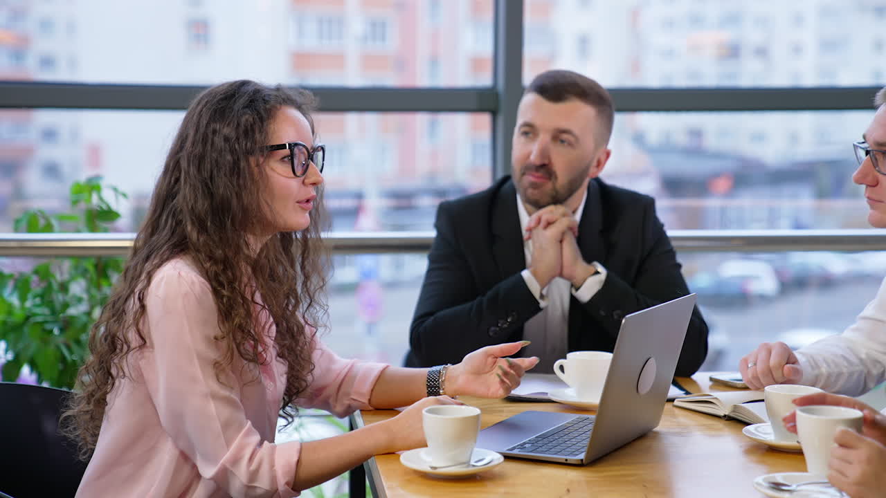 Beautiful curly-haired lady in glasses sits in front of laptop and speaks to her partners. Colleagues talk about professional issues at coffee time.