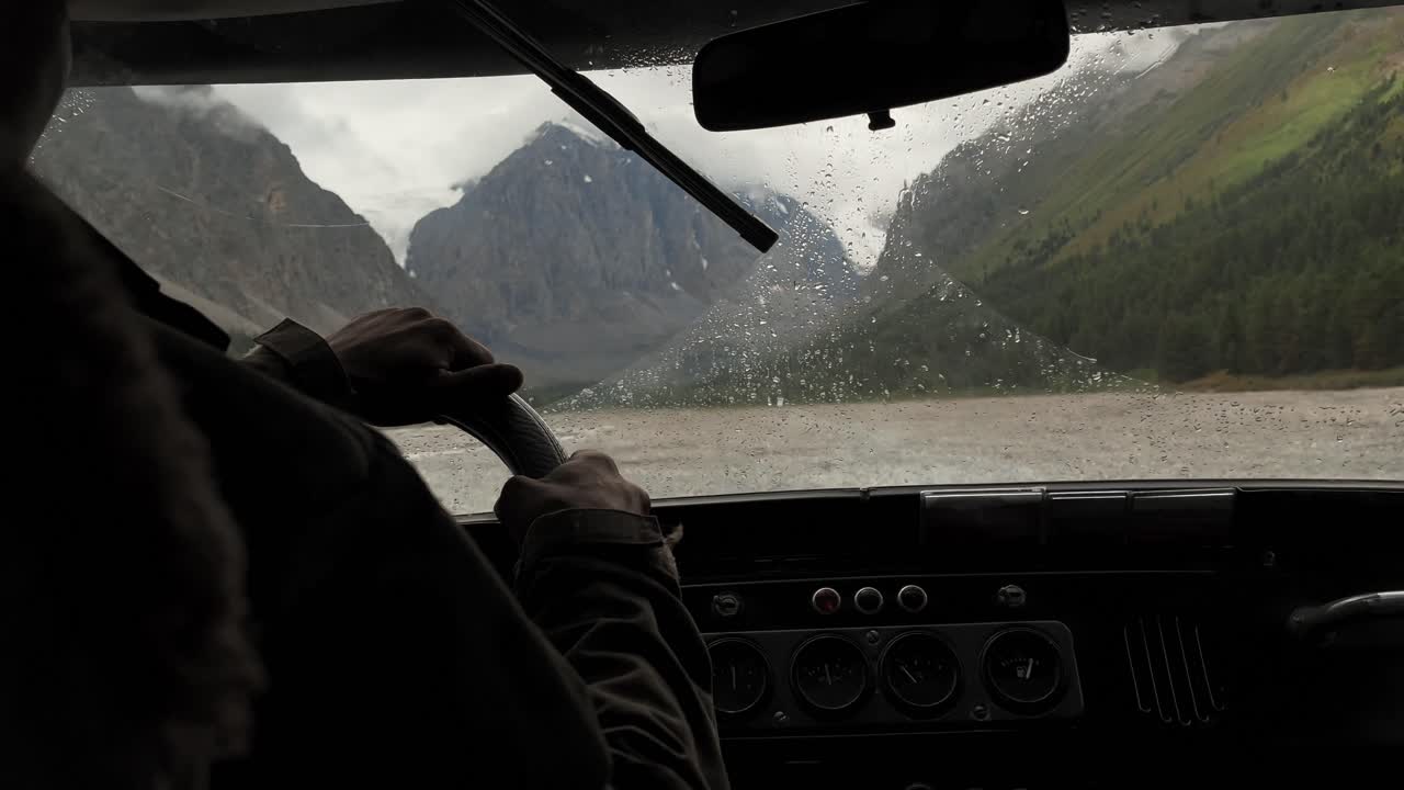 Point of view from inside a car driving in rain, windshield wipers clearing drops as mountains and a river valley pass by in Altai Republic, Russia, travel road trip mood