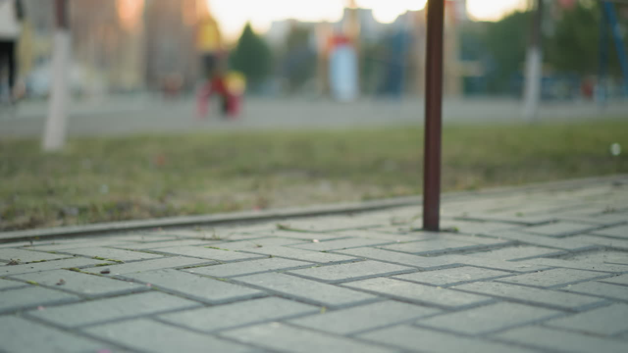 Close-up shot focusing on the intricate pattern of stone bricks on a paved park path. The background is subtle like a metal bench leg and hints of a park setting, including grass and distant figures