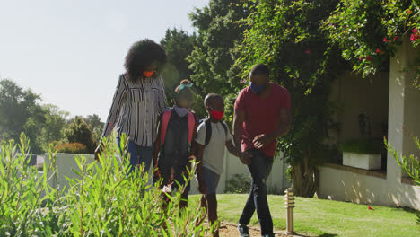 african american family wearing face mask walking in the garden holding hands together on a bright s