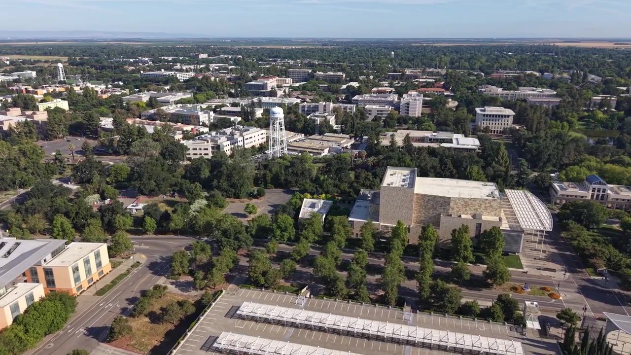 Vista aérea diurna con dron del campus de la Universidad de California, Davis, mostrando edificios académicos, estacionamientos con paneles solares, árboles verdes y la ciudad de Davis al fondo.