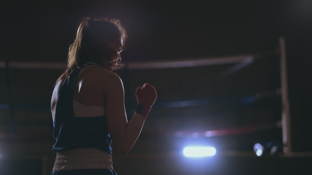 A beautiful woman conducts a shadow fight while practicing hard for future victories. Dark gym background. steadicam shot