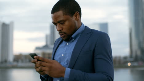 Businessman-In-Blue-Jacket-Standing-Near-River-And-Office-Buildings,-Holding-Cellphone-And-Chatting-With-Someone