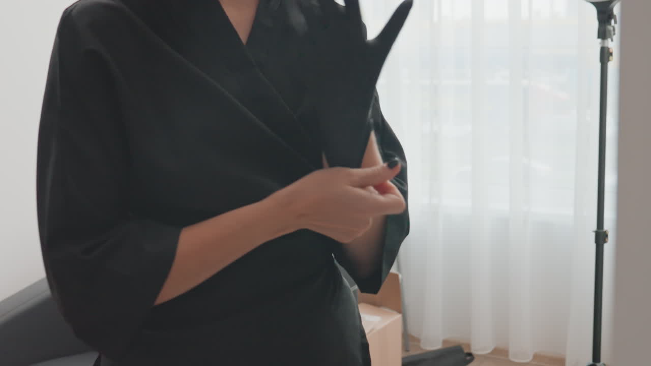 Donna in accappatoio che prepara i guanti, sistema lo scialle, allaccia il polsino, indossa un guanto nero in un luminoso studio spa con tende leggere, luce naturale soffusa, treppiede e sedia visibili, atteggiamento calmo e concentrato