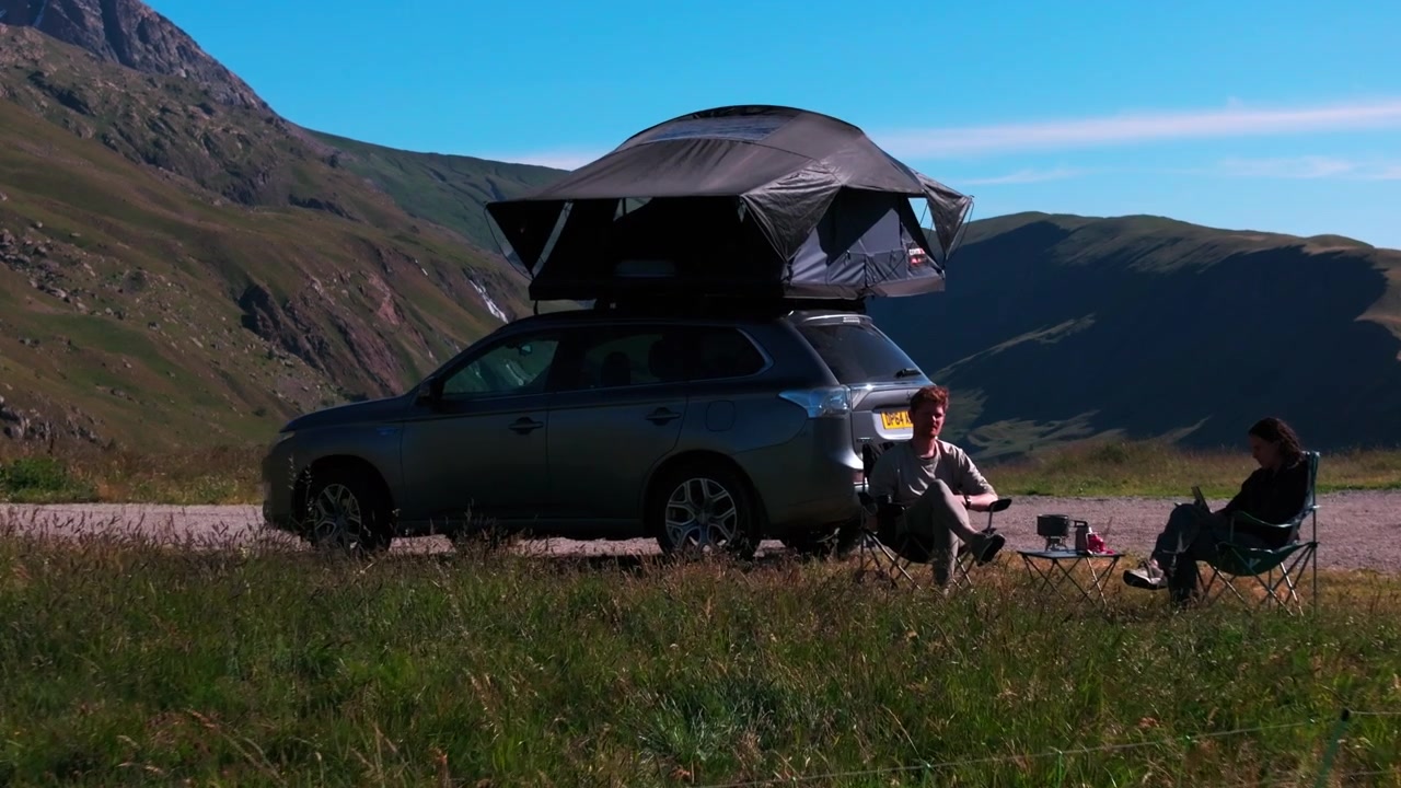 Un SUV con tenda da tetto è parcheggiato in una valle di montagna erbosa mentre una persona si rilassa accanto ad esso con un cane, incorniciato da cime distanti