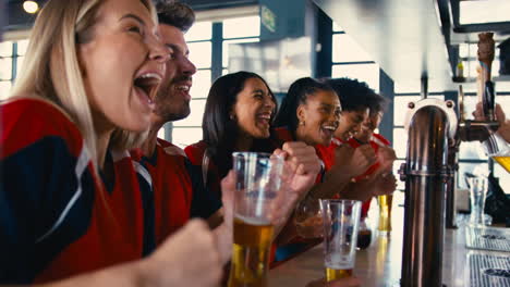 multi-cultural group of friends wearing team shirts in sports bar celebrating watching game on tv