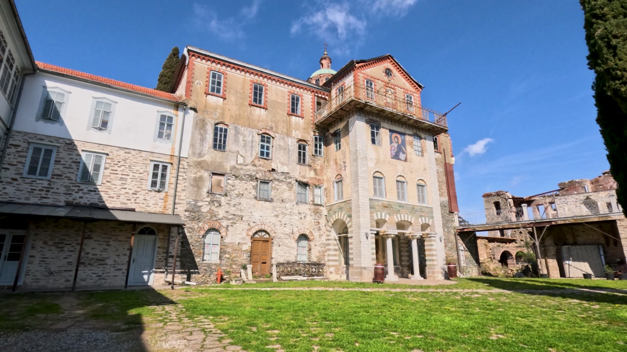 Visitors walk around an old monastery in Greece. The buildings show history with weathered walls and beautiful architecture. Sunlight shines on the stone path and grass area.