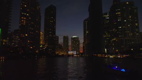 Low-forwards-fly-above-water-surface-between-silhouettes-on-high-rise-downtown-buildings-on-banks.-Night-city-scene.-Miami,-USA
