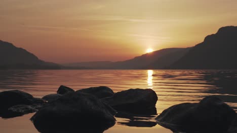 a dramatic shot of the sun setting behind a mountain which is casting a reflection on a  lake in ullswater, the lake district