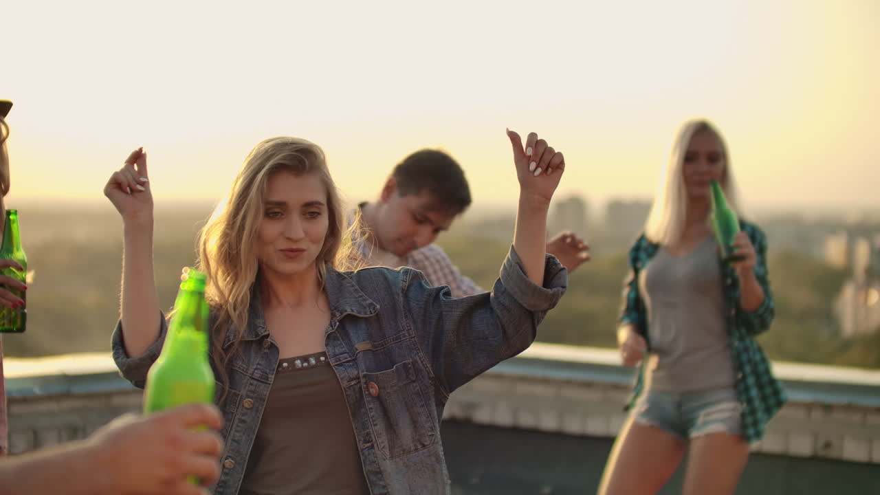 молодая девушка танцует на крыше со своими друзьями на вечеринке. она закрывает глаза и наслаждается временем в шортах и легкой джинсовой куртке в летнее вечное время. ее светлые волосы летают на ветру. она танцует руками и телом.