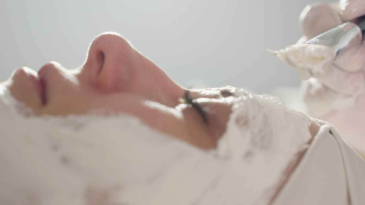 Hands of aesthetician spreading creamy face mask in circular motions with silicone brush over resting model s forehead cheeks chin under soft studio lighting conveying calm spa treatment