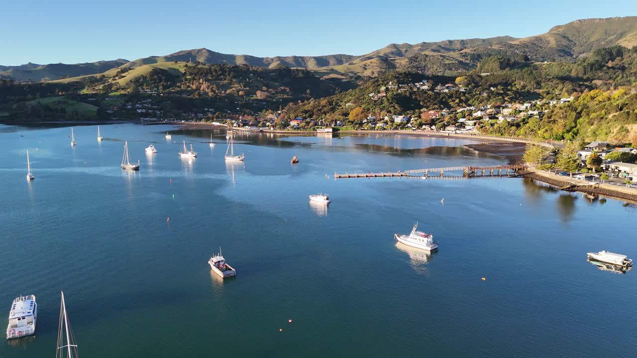 Drone footage captures serene Akaroa harbor with boats, calm waters, and scenic hills during sunset in Canterbury, New Zealand