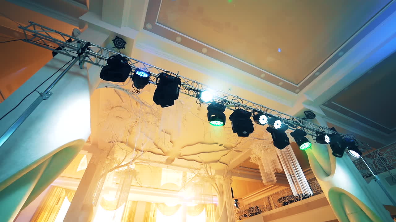 Black projectors hanging from top in the light concert hall. Metal construction with colorful spotlights on the creamy background indoors.