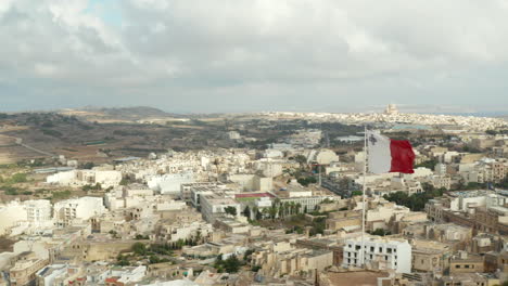 Flagge-Von-Malta-Weht-Im-Wind-Mit-Blick-Auf-Die-Stadt-Auf-Der-Insel-Gozo,-Antenne-Nah-Oben-Schieben-Nach-Links