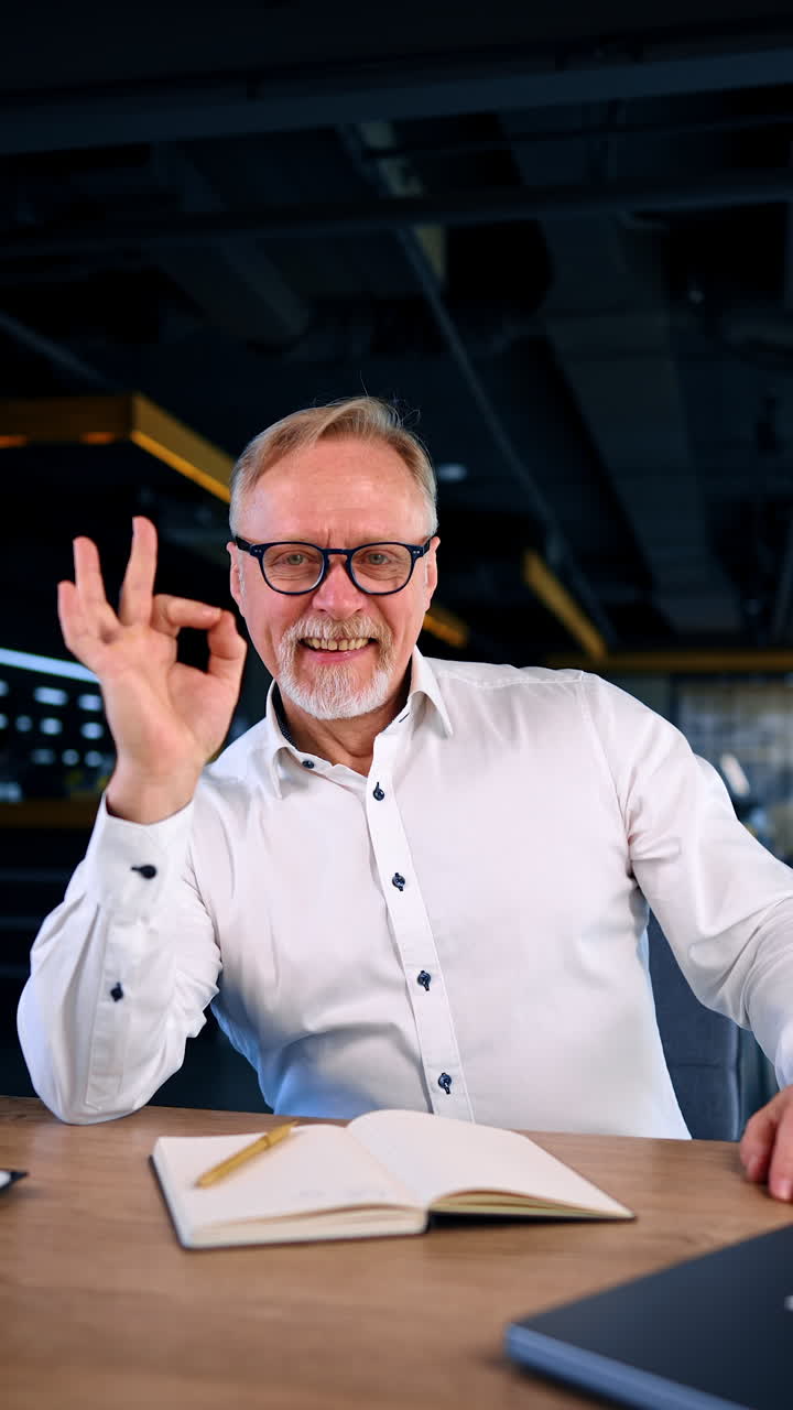 Positive Caucasian male in white shirt sits at desk in the office. Cheerful man gestures to camera. Vertical video.