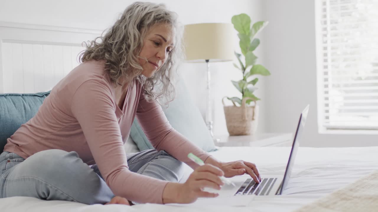 Happy senior caucasian woman sitting on bed and using laptop, slow motion