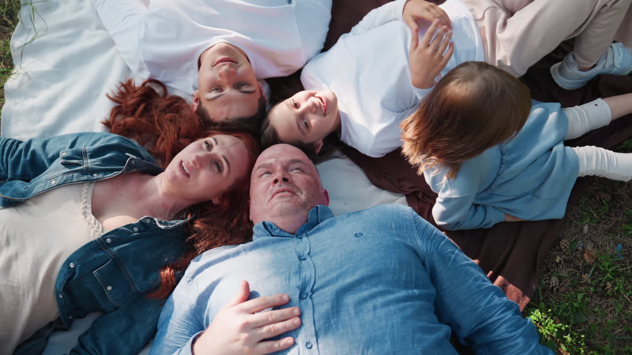 Aerial view of nature loving family lying down on blanket with heads joined, talking and smiling warmly under sunlight as youngest joins them, showing togetherness, happiness, and peaceful bonding