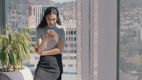 Hermosa-Mujer-De-Negocios-Usando-Un-Teléfono-Inteligente-Enviando-Mensajes-De-Texto-A-Una-Ejecutiva-Revisando-Correos-Electrónicos-Explorando-Mensajes-En-Un-Teléfono-Móvil-En-La-Oficina-Parada-Junto-A-La-Ventana-Con-Vista-A-La-Ciudad