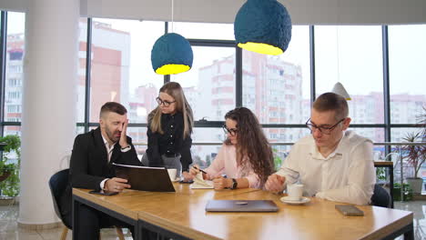 tired boss sits at the head of the table talking to his staff. female employees listen to their chief and man drinks coffee.