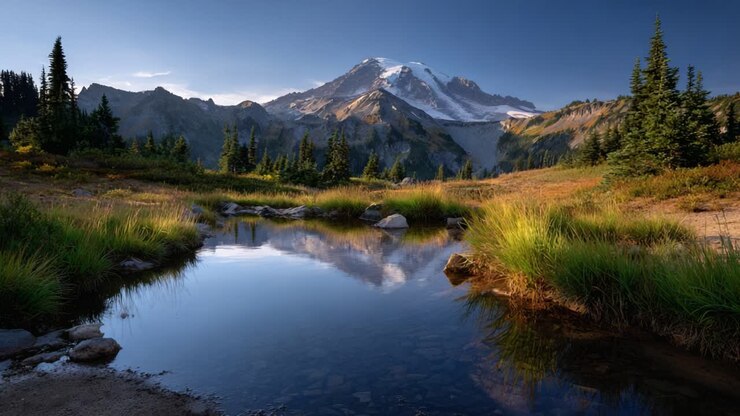 Paesaggio montano mozzafiato con riflessi in un tranquillo stagno, circondato da vegetazione lussureggiante e vette maestose, sotto un cielo azzurro limpido al tramonto.