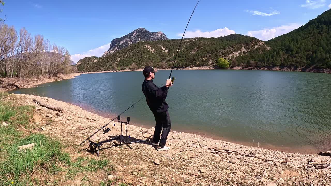 A young man stands on a rocky lake shore, skillfully reeling in a fish with his fishing rod. The carp fishing setup, including a tripod, is visible. Scenic mountainous landscape