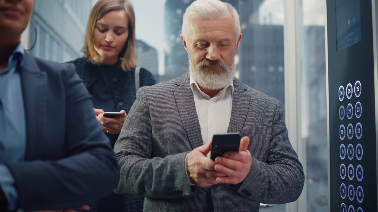 Successful Middle Aged Businessman Riding Crowded Glass Elevator to Office in Modern Business Center. Handsome Happy Man Using Smartphone, Write a Message, Social Media and Work Emails in a Lift.
