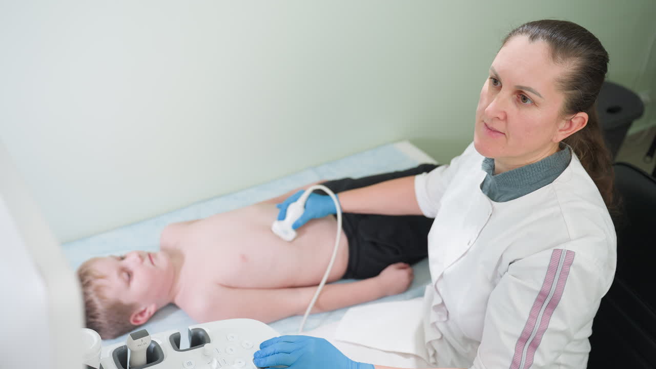 Female lab technologist seated with focused gaze on examination monitor while using scanner on barechested teenager lying on examination bed during diagnostic ultrasound in clean clinical environment