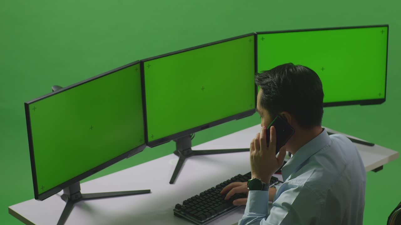 녹색 스크린 스튜디오에서 여러 컴퓨터 모니터를 모방하여 작업하는 동안 휴대 전화로 대화하는 아시아 남자의 측면 시각